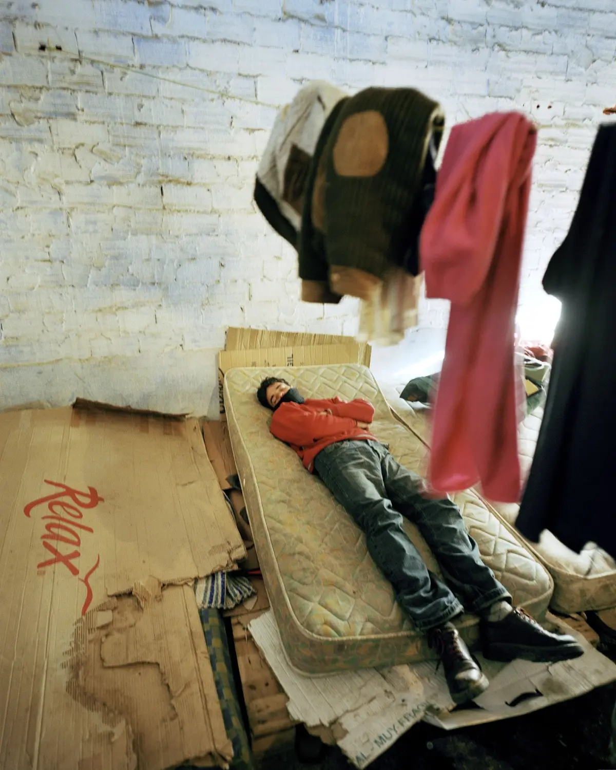 Person sleeping on a mattress in a cluttered room with hanging clothes and a rough brick wall.
