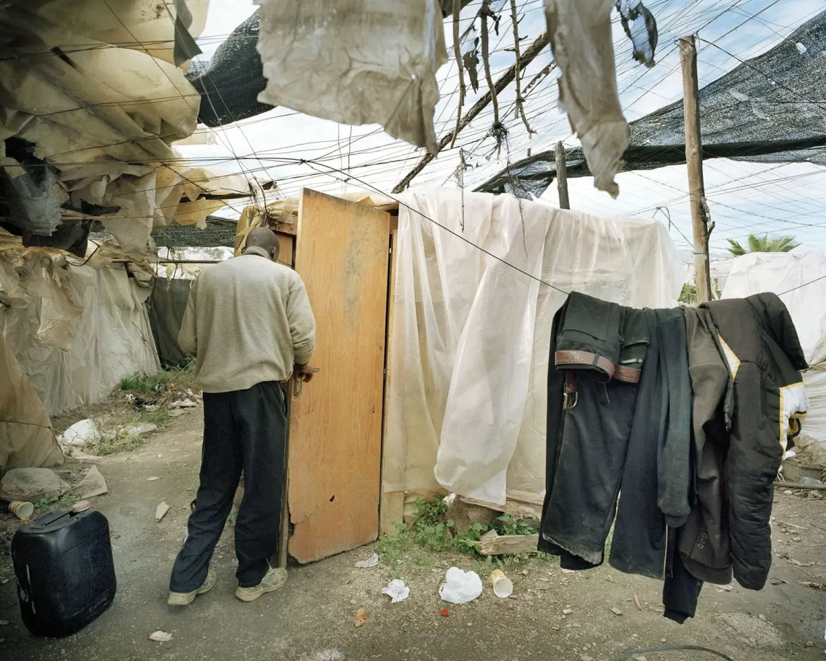 Person in gray hoodie stands by an open wooden door inside a tarp-covered makeshift shelter; clothes hang on a line nearby.