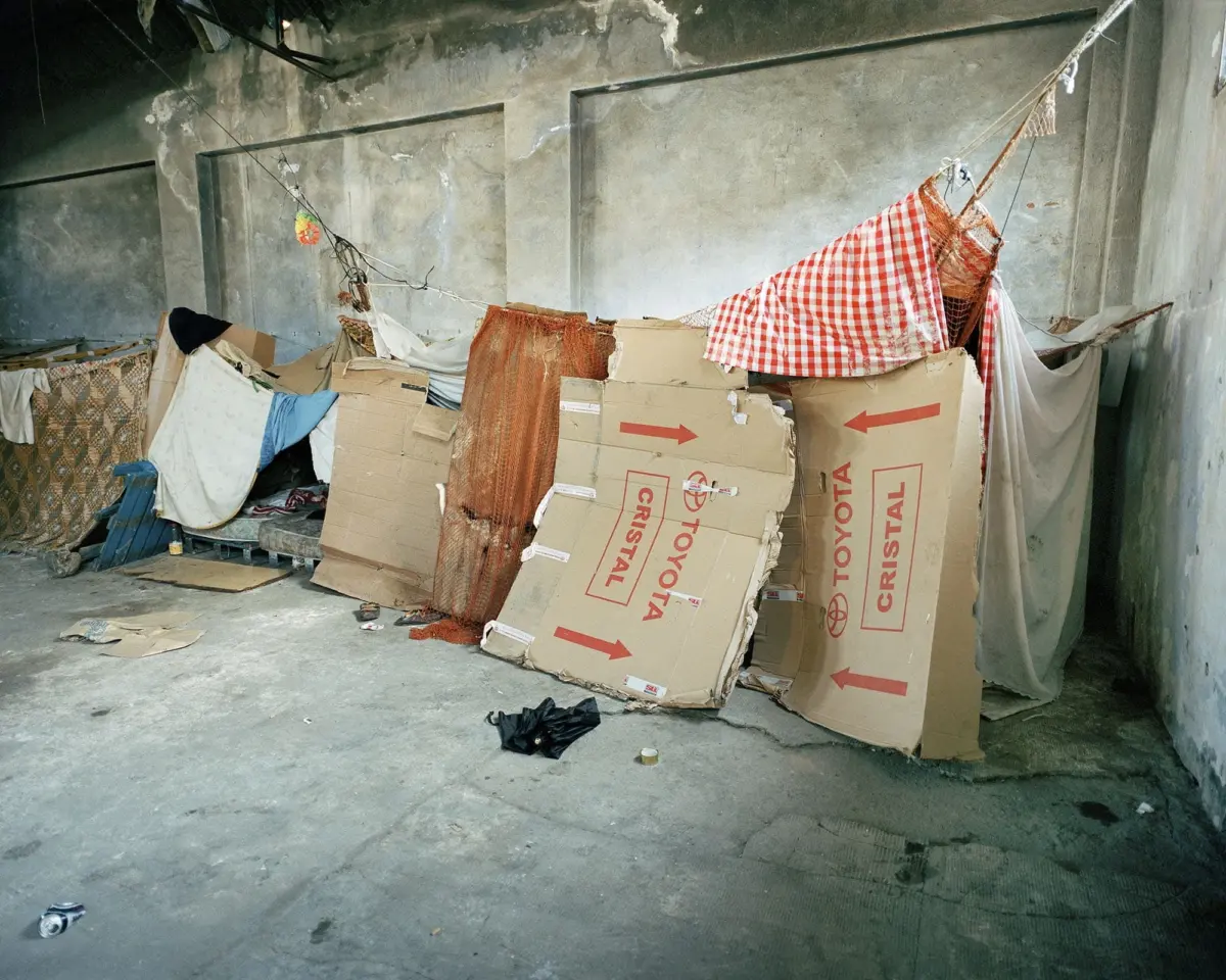 Makeshift encampment of cardboard boxes and fabric under a concrete underpass, with a clothesline and red-checkered sheets.