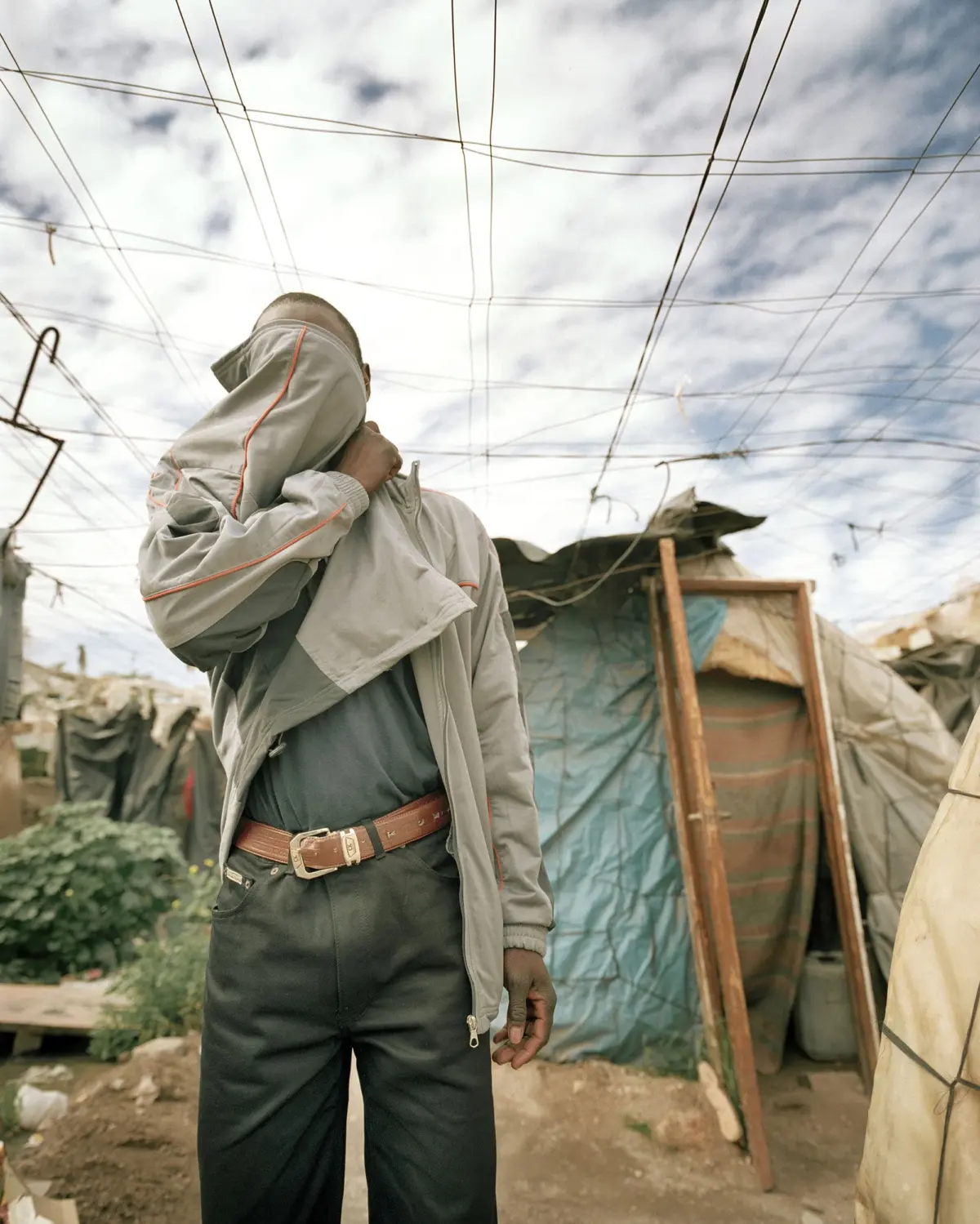 Person in gray jacket and dark pants stands in a makeshift camp with tarpaulin tents, lifting the jacket to cover the face amid overhead wires.