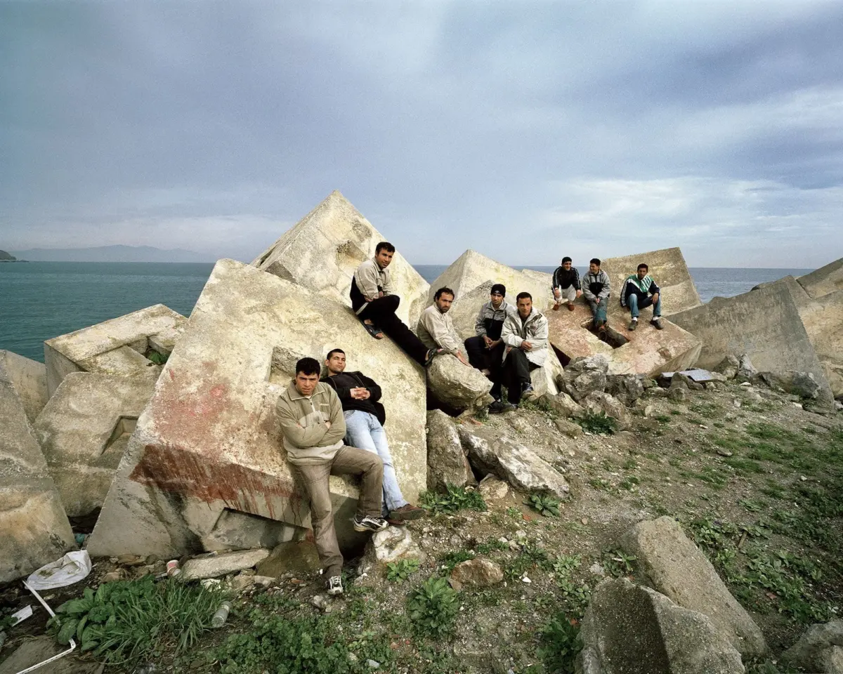 Group of men seated on large concrete tetrapod blocks along a rocky coastline by the sea.