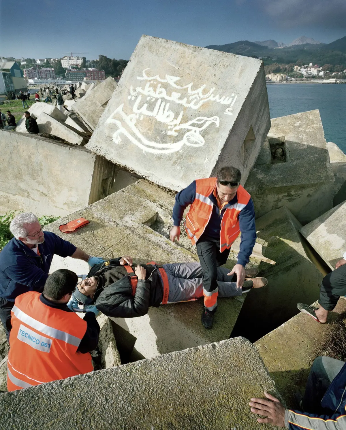 Rescue workers in orange vests carry an injured person on a stretcher along a rocky breakwater by the sea, with a cityscape in the distance.