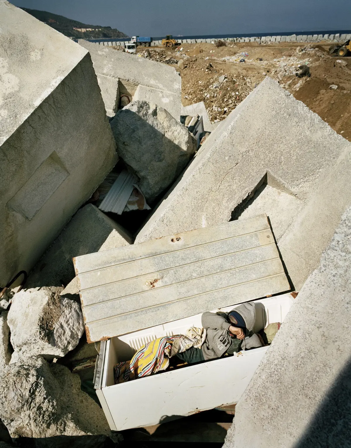 Person lying inside an open refrigerator amid large concrete blocks at a coastal construction site, with distant machinery and seawall visible.