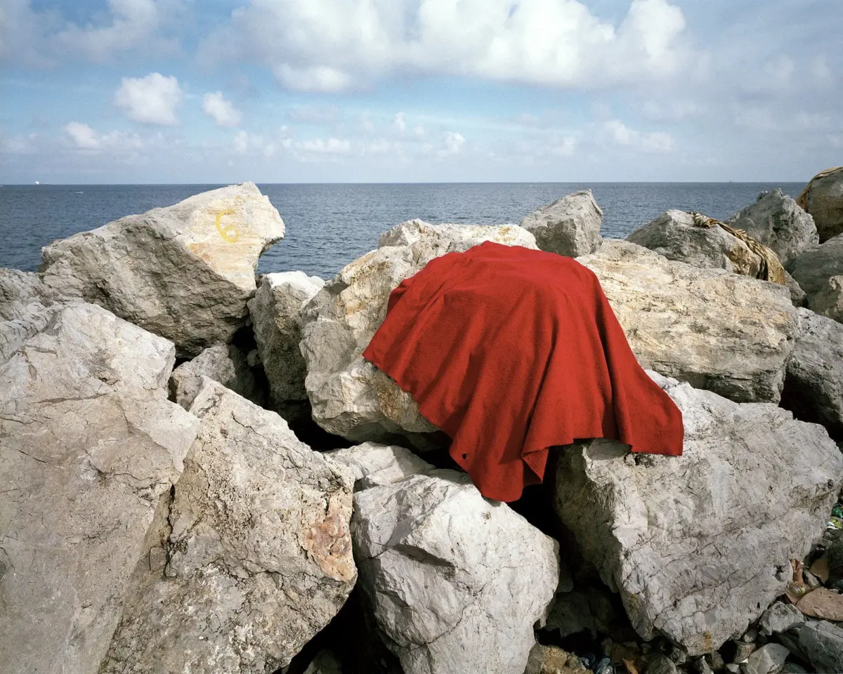 Red cloth draped over gray rocks along a rocky shoreline, with the sea and a cloudy blue sky in the background.