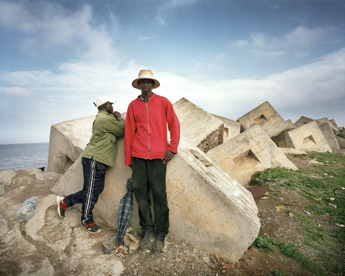 Two people stand among large concrete blocks by the sea; one in a red jacket stands upright, the other in green leans on a block.