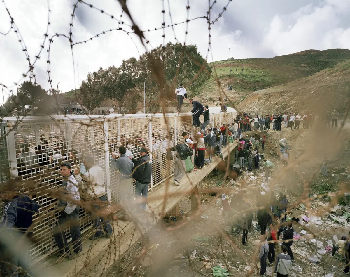 Large crowd along a barbed-wire fence on a pedestrian bridge over a rocky hillside; some individuals climb or balance on the railing.