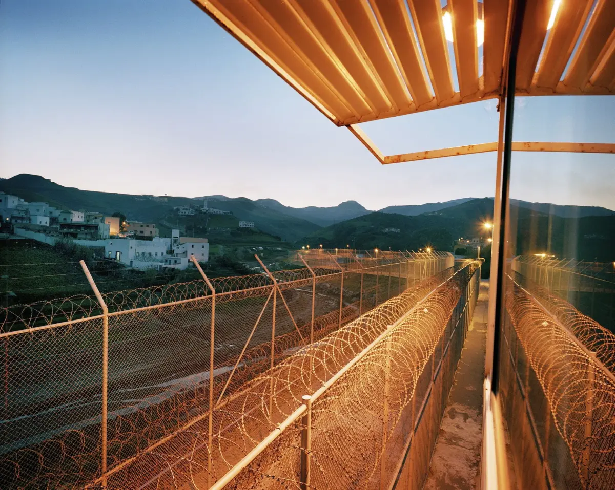 Barbed-wire fence along a concrete walkway beside a glass building at dusk, with a hillside town in the distance.