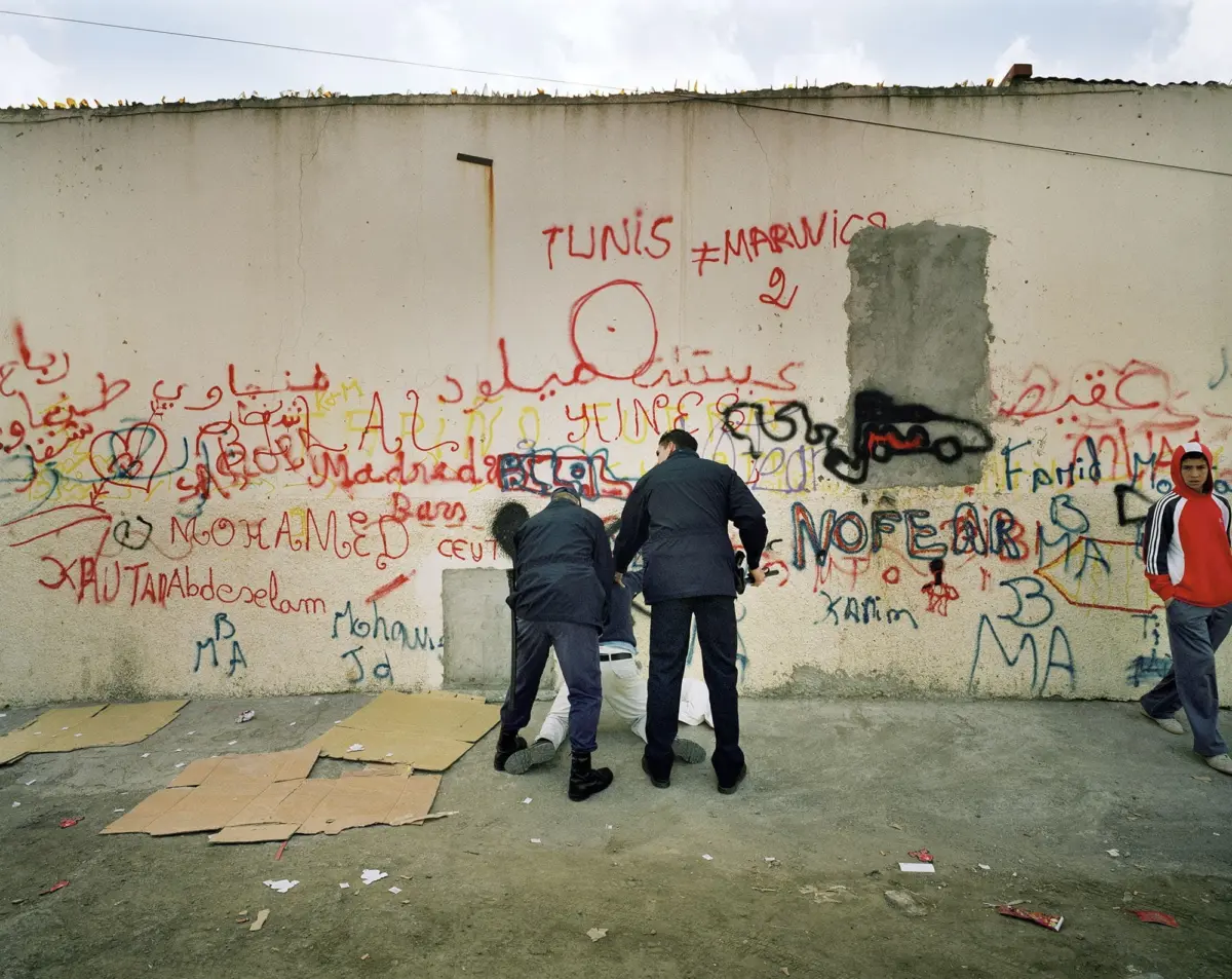 Group of people standing and crouching by a graffiti-covered wall with large red text 'TUNIS'; cardboard sheets scattered on the ground.