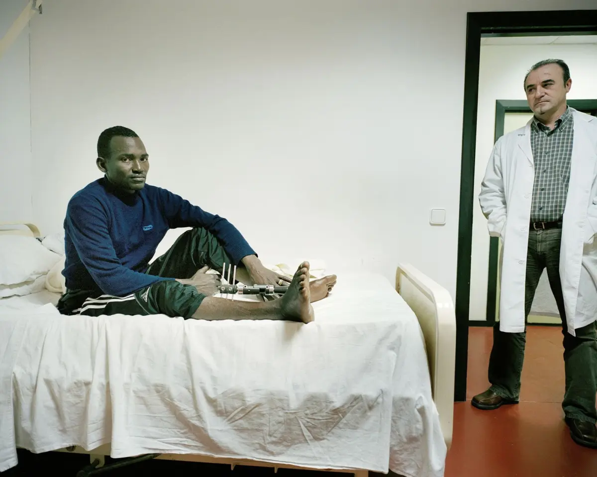 A person sits on a hospital bed with a prosthetic leg, in a hospital room, as a doctor in a white coat stands in the doorway.