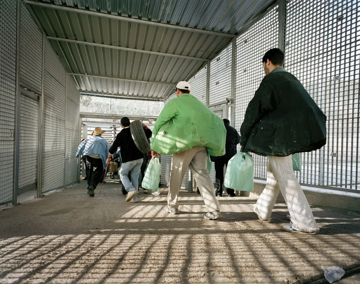 Group of people walking through a metal mesh corridor, carrying large green bags and a tire.