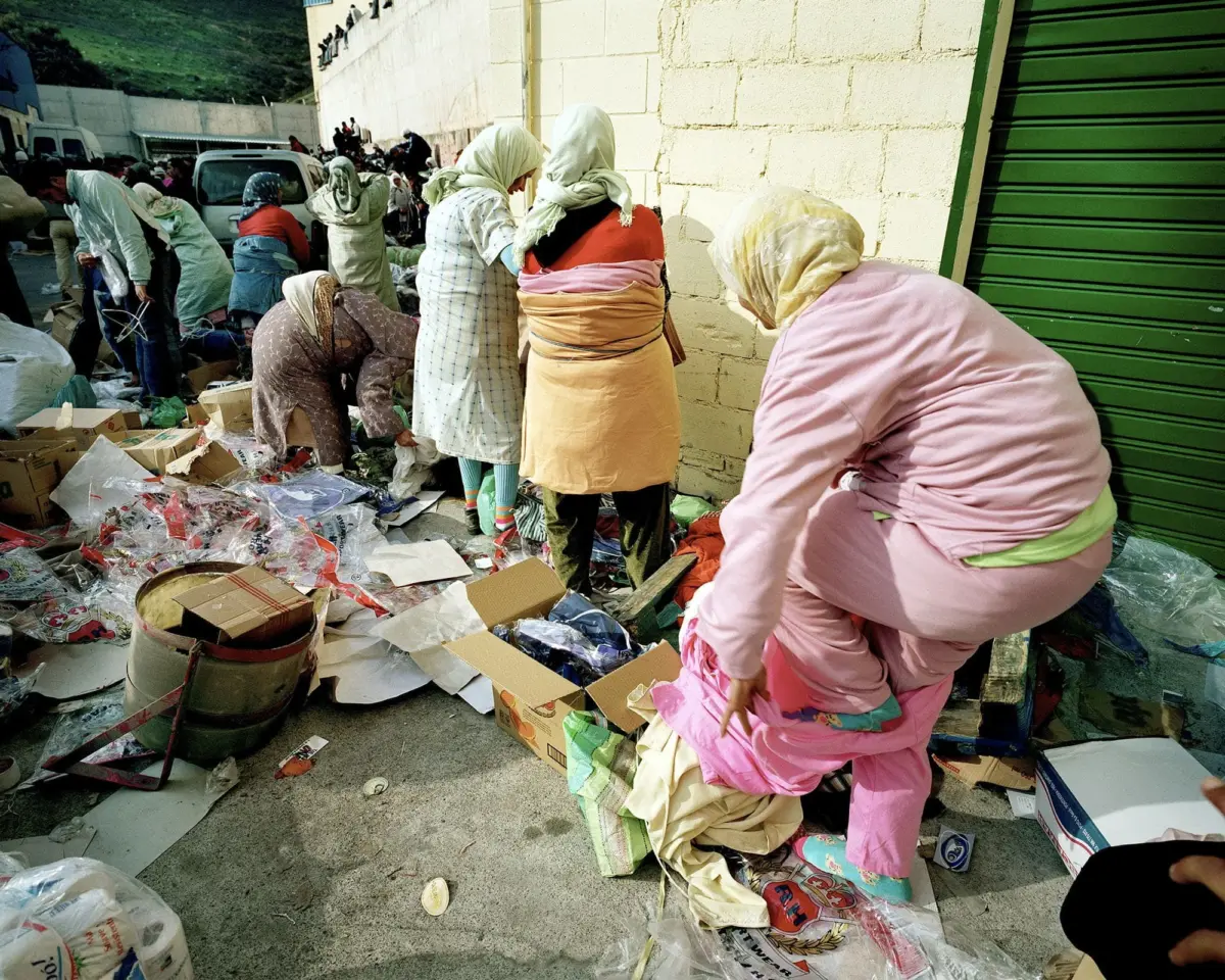 Group of women in headscarves rummaging through garbage and cardboard debris along a concrete street beside a beige brick wall
