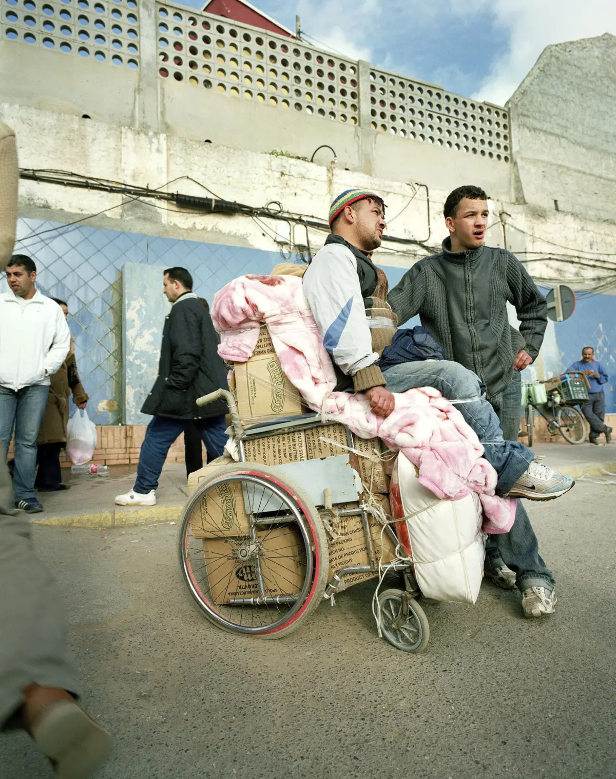 Two men on a makeshift cart loaded with cardboard boxes and pink blankets on a street; one sits, the other stands beside, with pedestrians nearby.