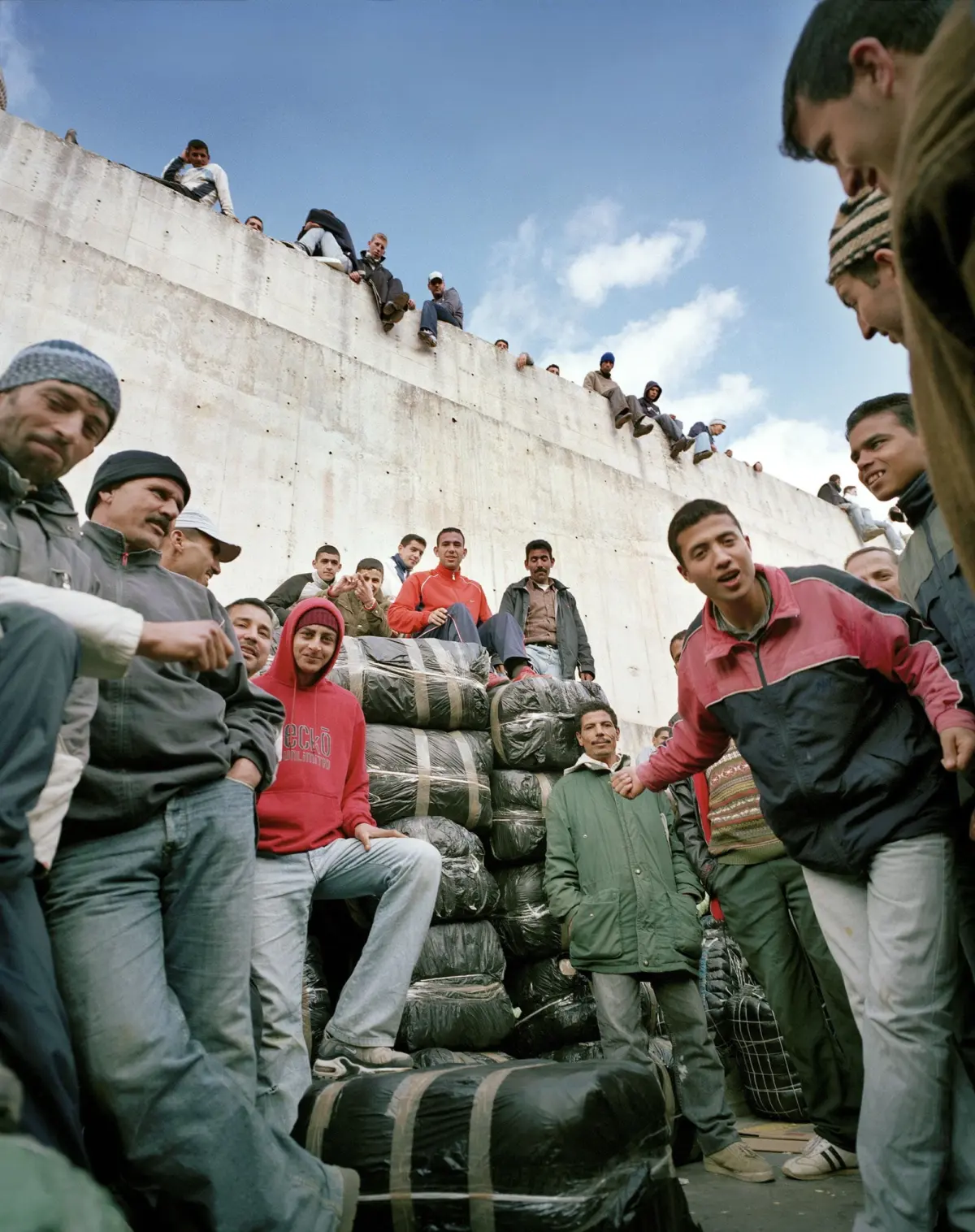 Group of people in jackets atop a concrete wall, looking down at cargo wrapped in black plastic below under a blue sky.