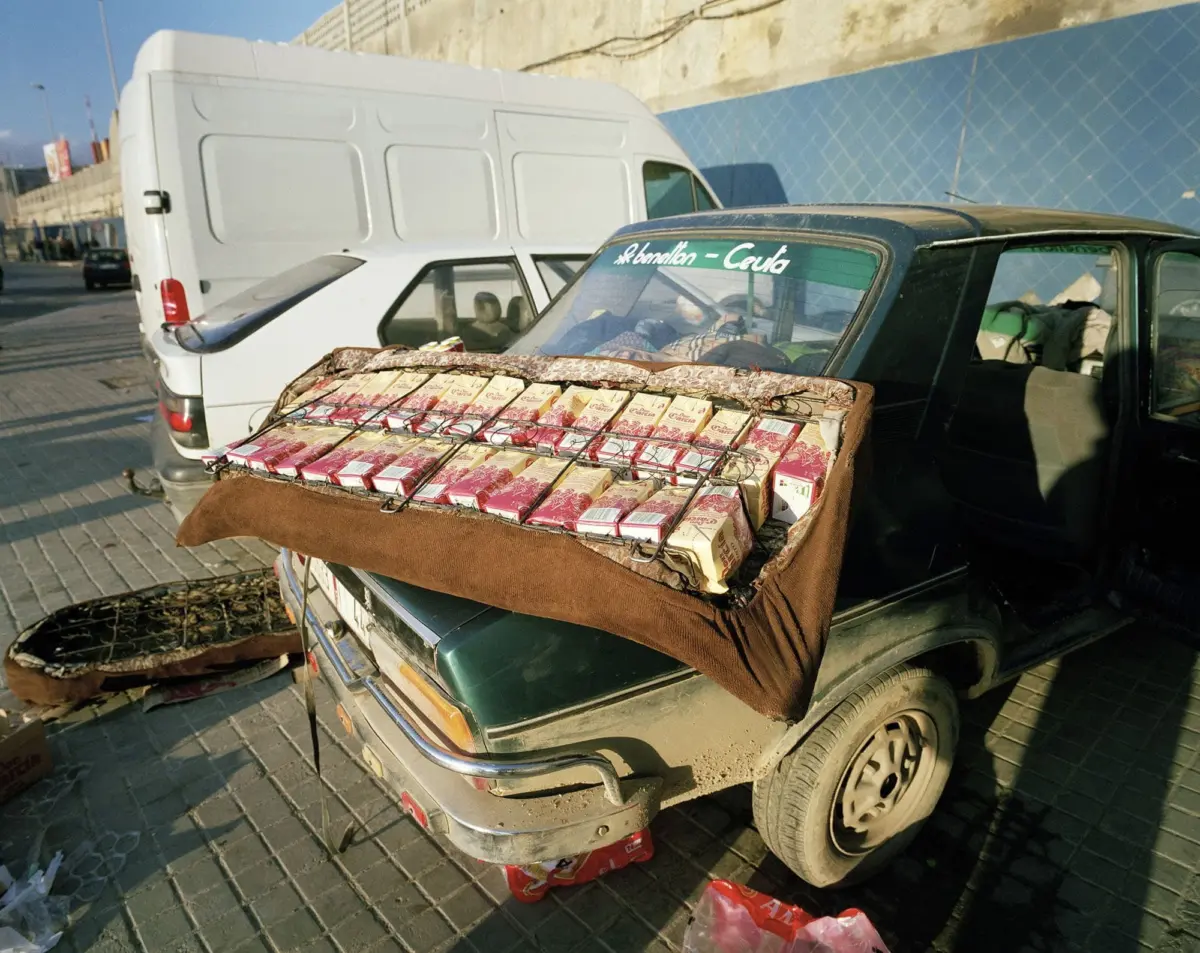Green SUV with a rack of cigarette packs laid on its hood, parked on a city street beside a white cargo van.