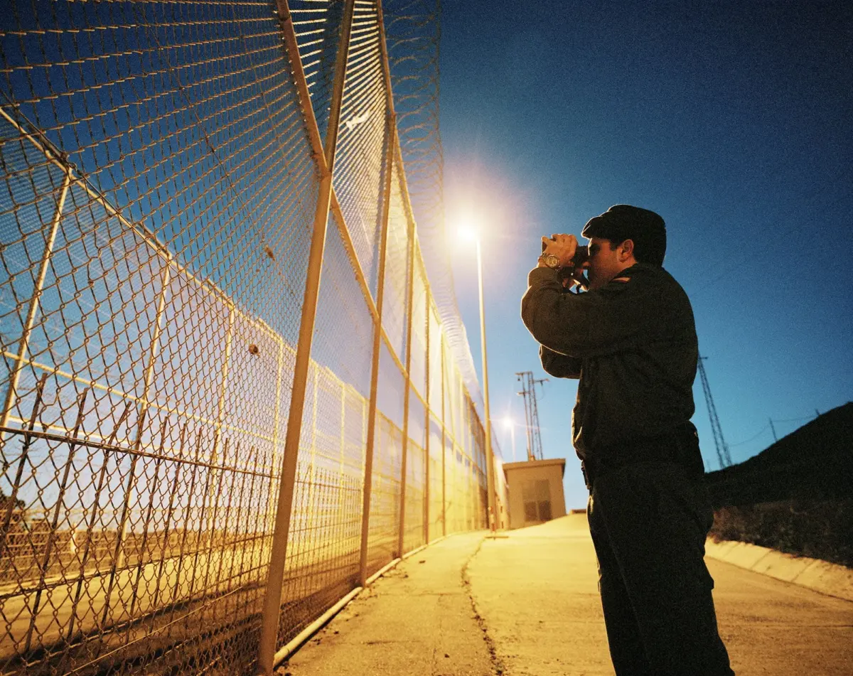 Photographer takes a nighttime photo along a chain-link fence beside a lit industrial area.