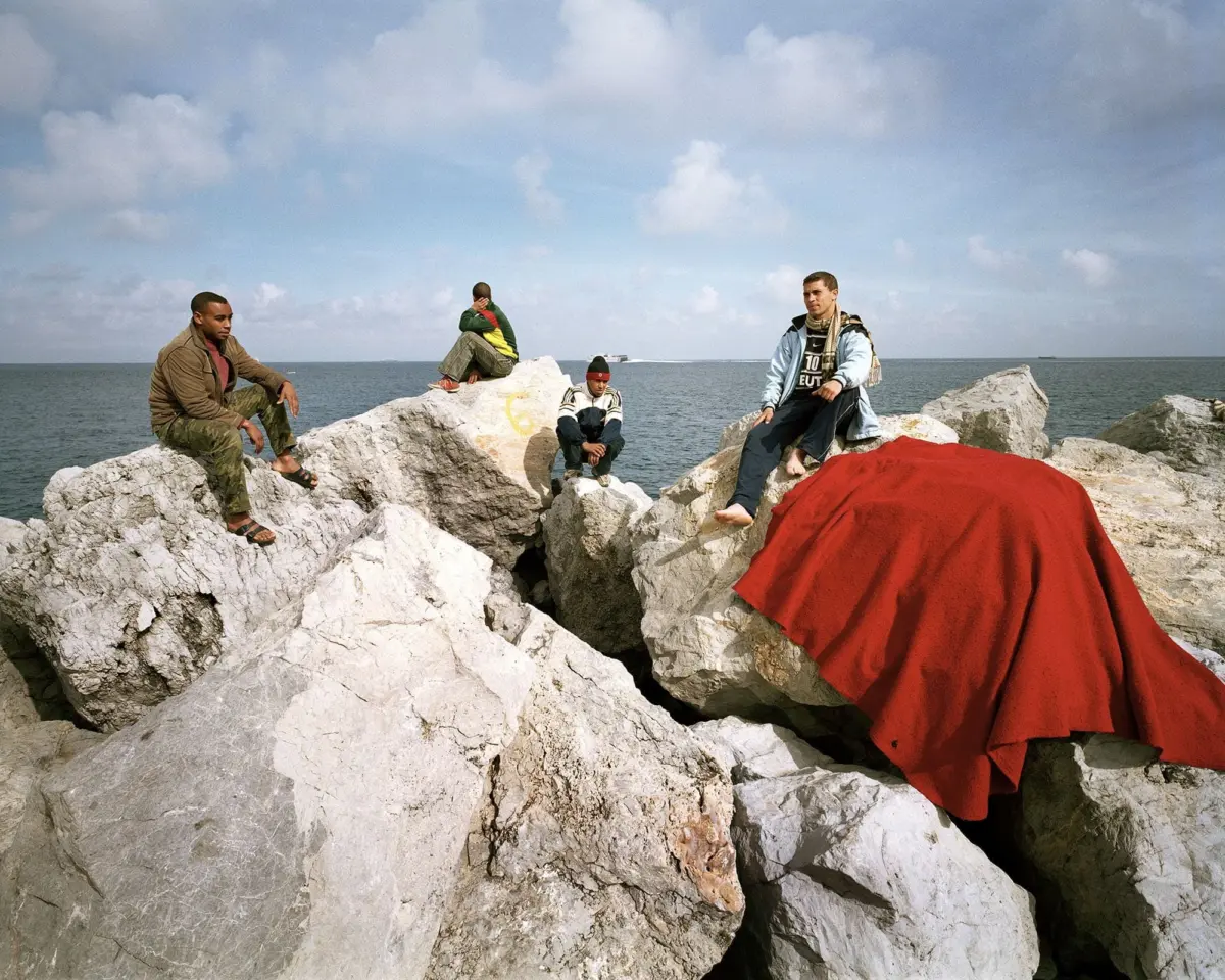 Four men sit on jagged white rocks by the sea under a cloudy sky, with a large red fabric draped in the foreground.