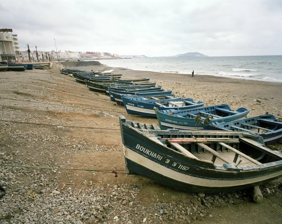 Blue fishing boats lined up on a pebbled beach with a man standing near the surf; distant beachfront city under cloudy sky.