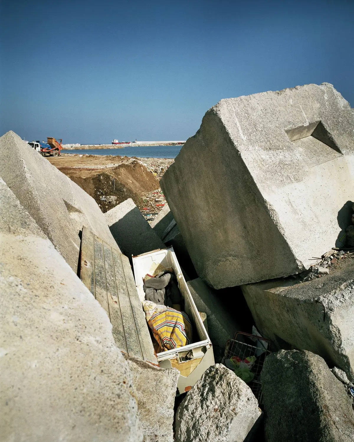 Human figure lying inside a damaged cabinet amid rubble and large concrete blocks on a beach.