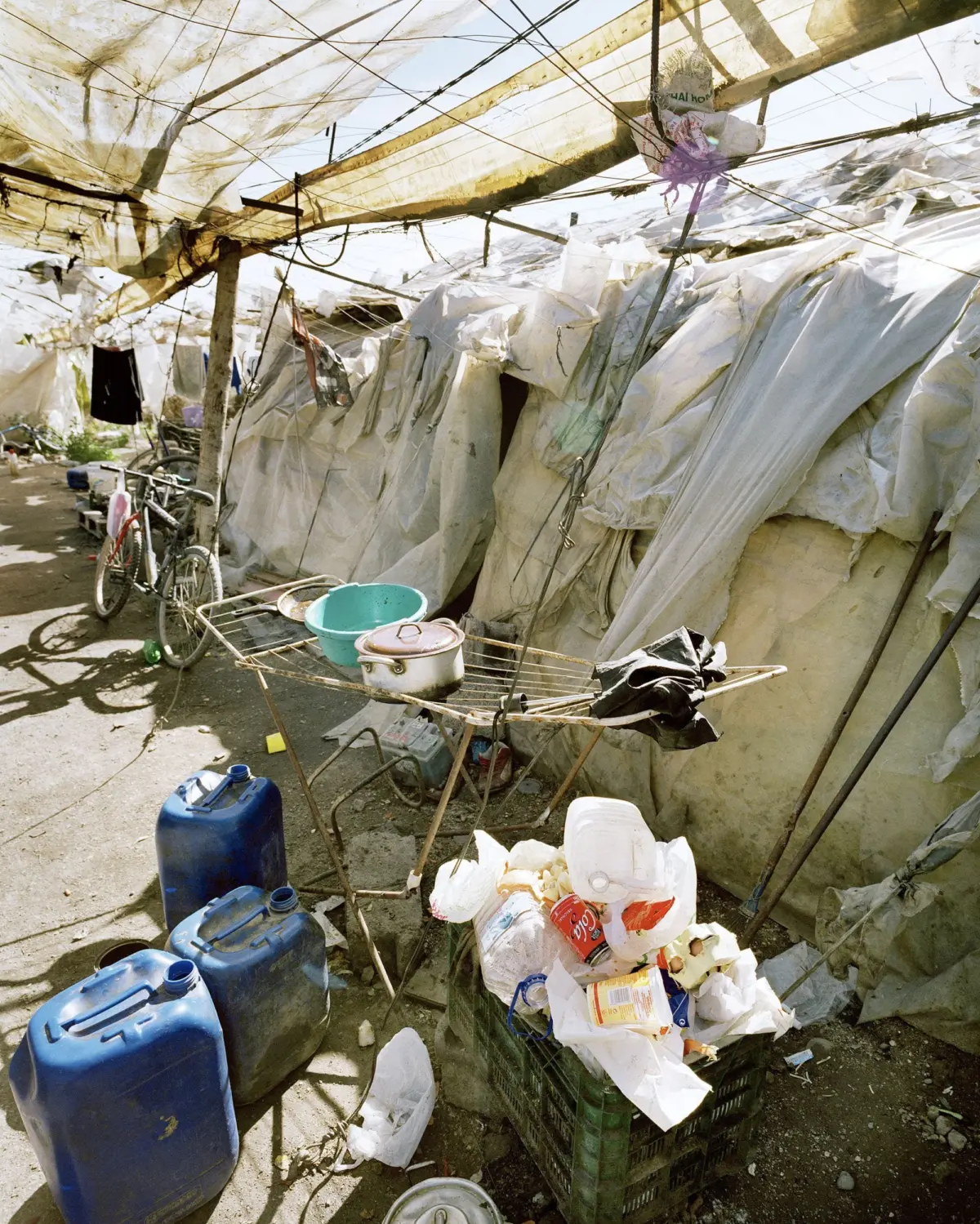 Makeshift outdoor camp with plastic-sheet shelters; cluttered cooking area on a metal rack with a pot and kettle, near blue jerrycans and a bicycle.