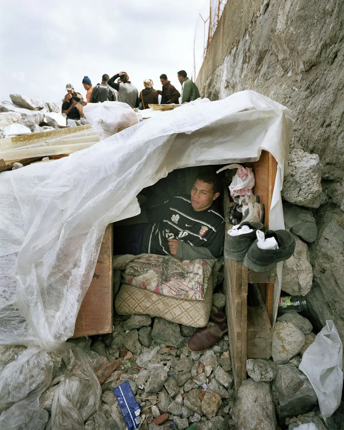 Group of people on a rocky shoreline near a tarp-covered wooden shelter; a man sits inside while others chat.