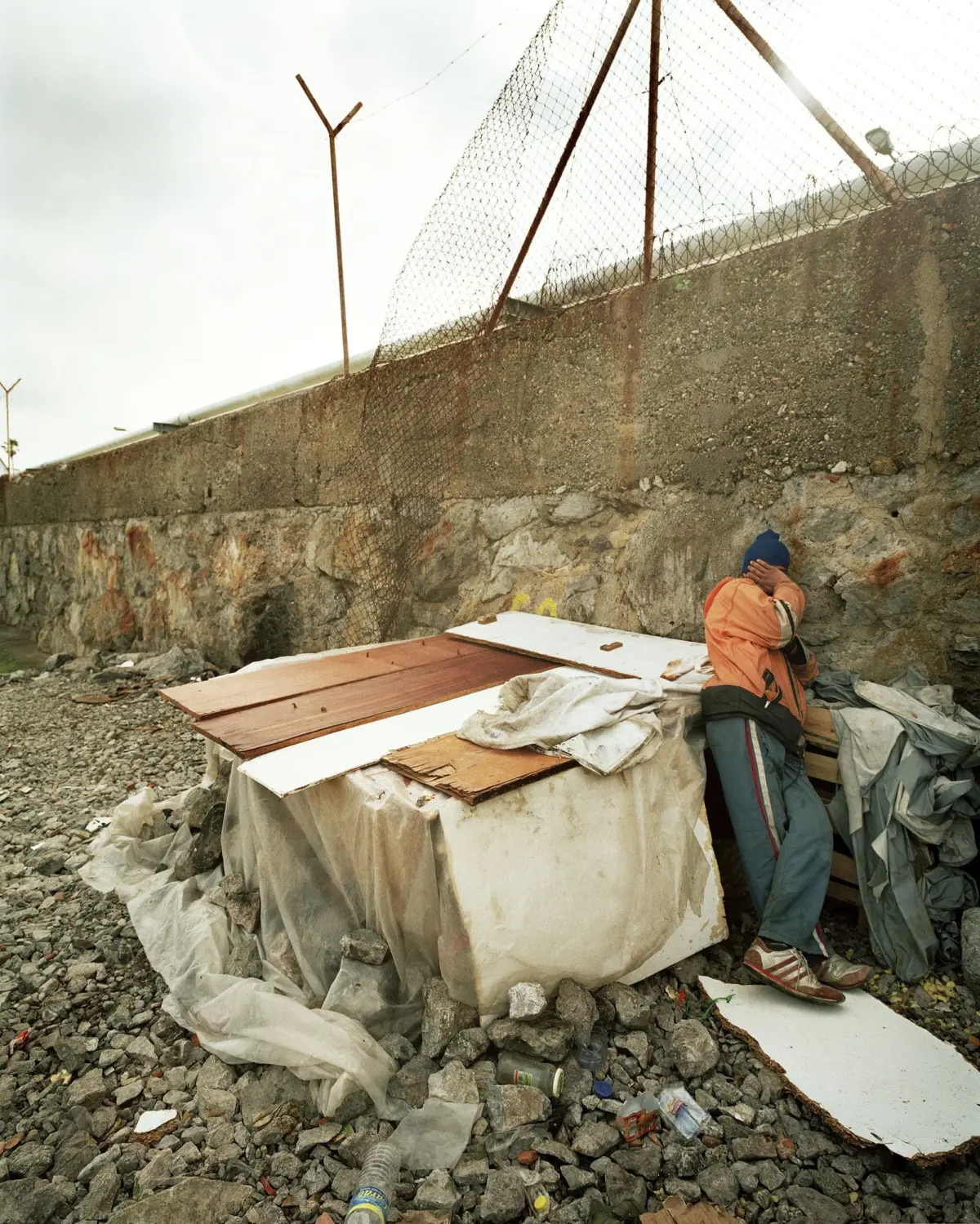 Person in orange jacket and blue beanie covers their face while leaning on a tarp-covered rubble stack beside a stone wall topped with barbed-wire fence.