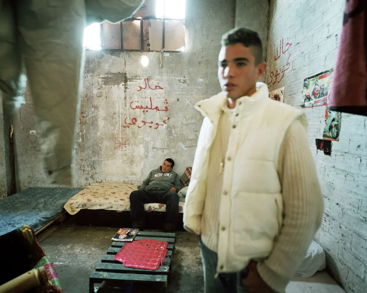 Person in a light quilted jacket stands in a graffiti-covered, dilapidated room; a second person sits on a bed.