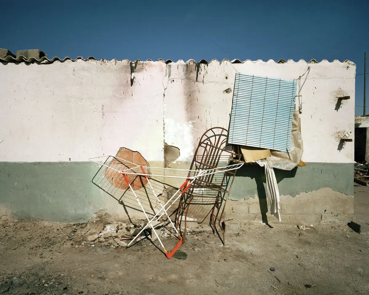 Discarded metal drying racks and bent chair frames leaning against a peeling white wall in a dusty, debris-strewn yard.