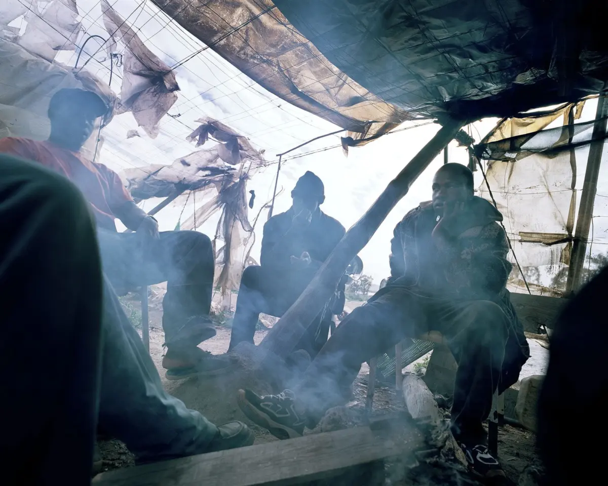 Three people sitting inside a makeshift shelter of torn plastic sheets and wooden poles, smoking amid hanging fabric and wires.