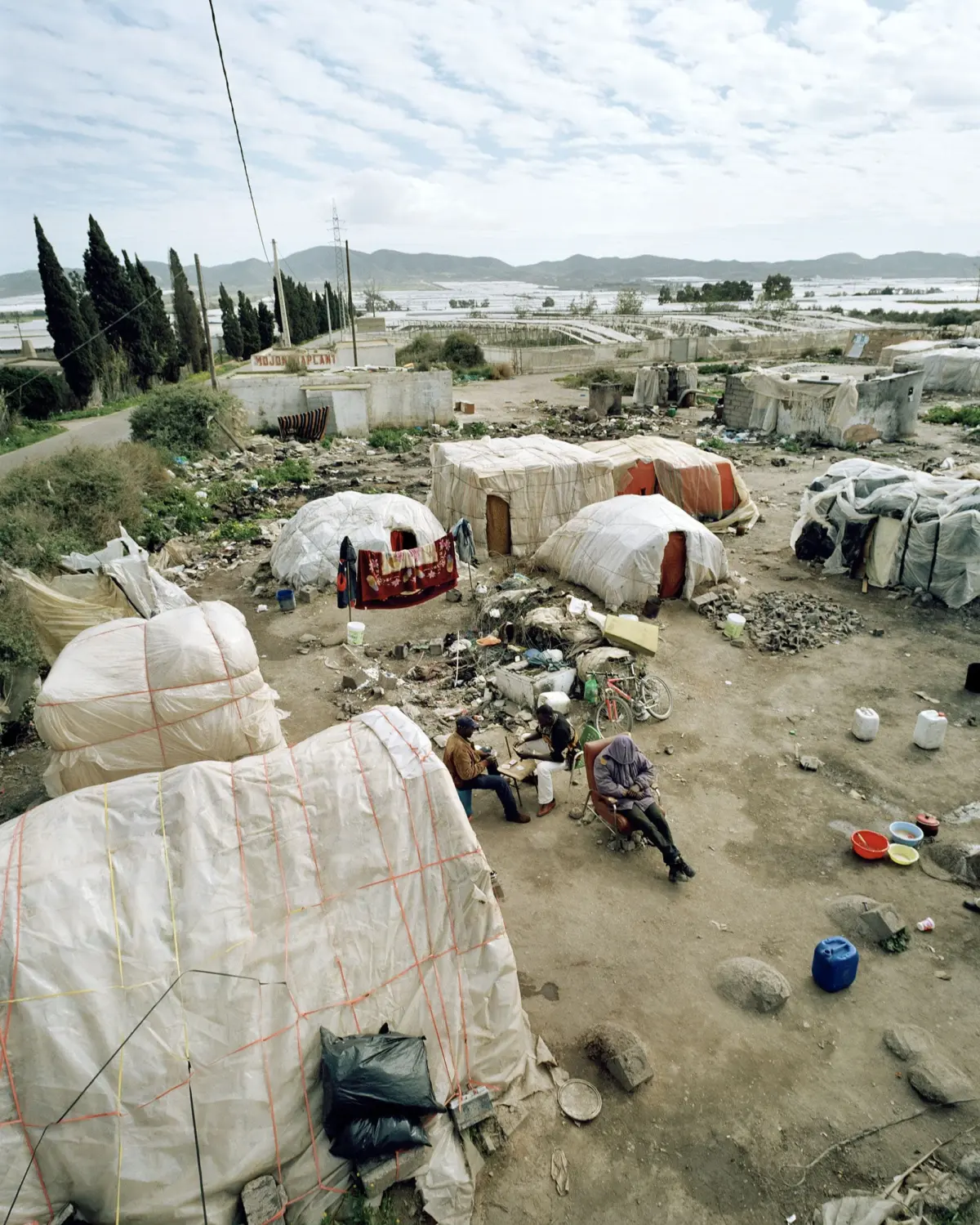 Desert refugee camp with white fabric tents tied by orange ropes; a few people sit around a table amid debris, distant hills in the background.