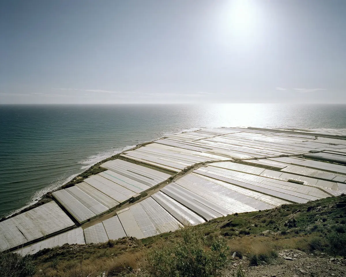 Coastal salt evaporation ponds covered with white plastic sheets arranged in a grid along a hillside by the sea.