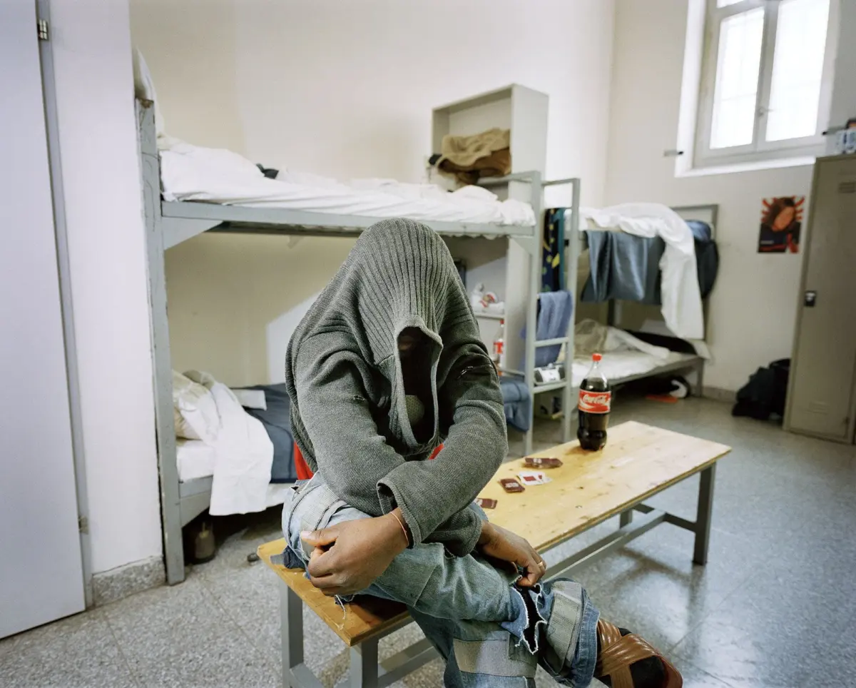 Person in gray hoodie sits on a wooden bench in a dorm room with bunk beds; face obscured by hood, arms folded, clutter around.