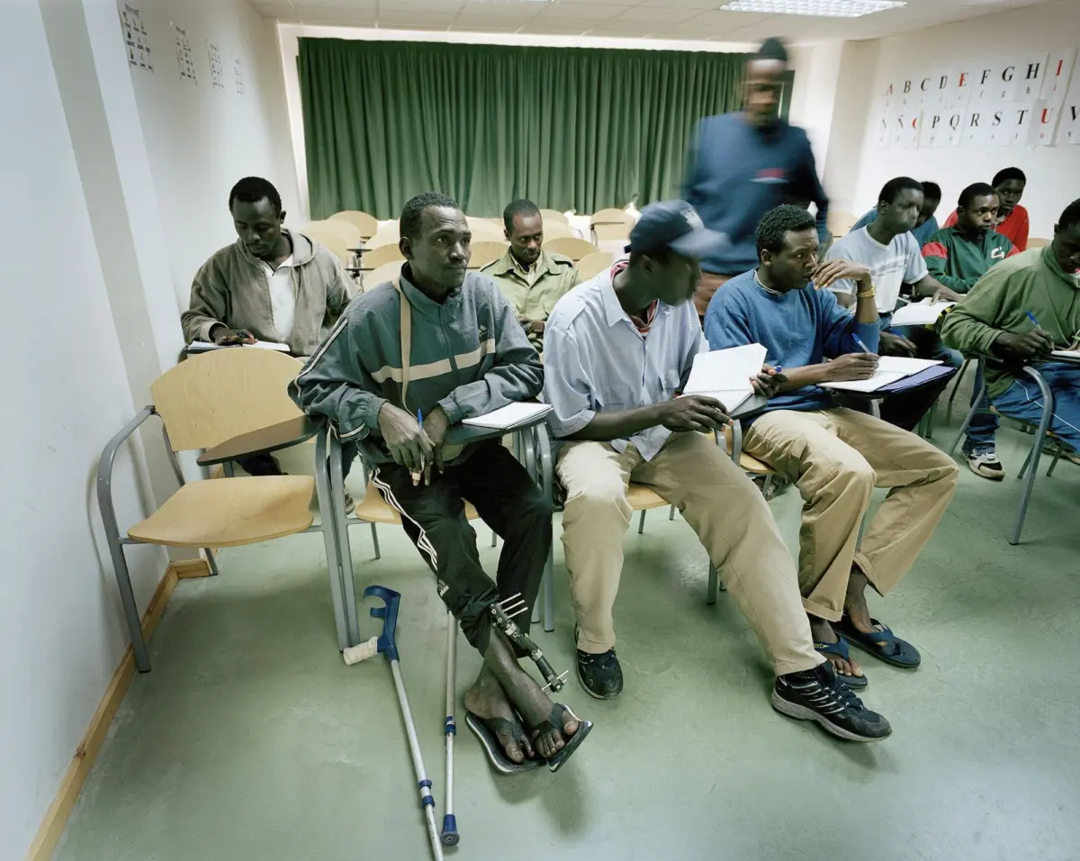 Group of men in a classroom; some write in notebooks, foreground man with leg prosthetics sits with crutches.