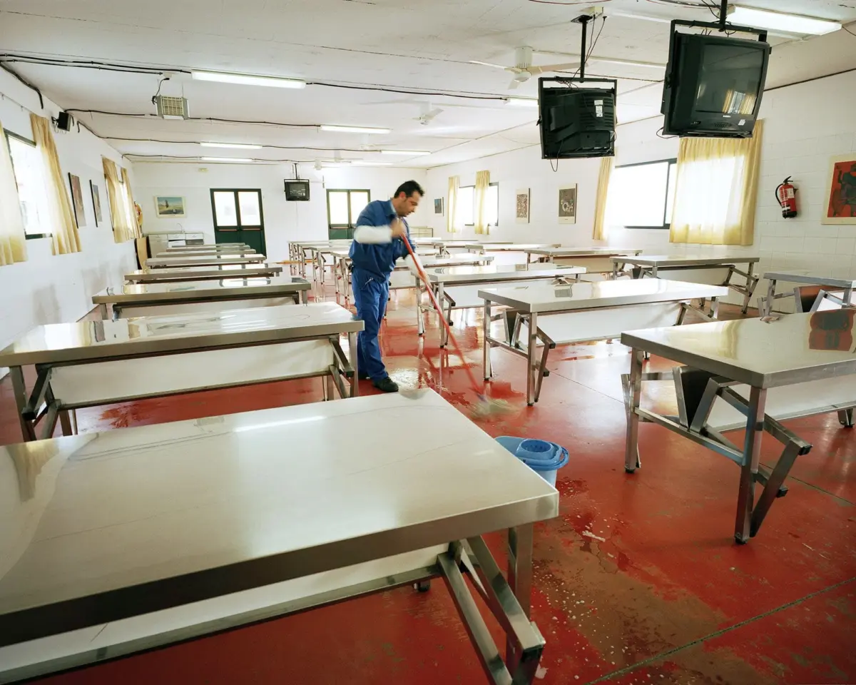 Janitor in blue uniform mops a wet red-floored cafeteria with stainless steel tables, ceiling-mounted TVs, and yellow-curtained windows.