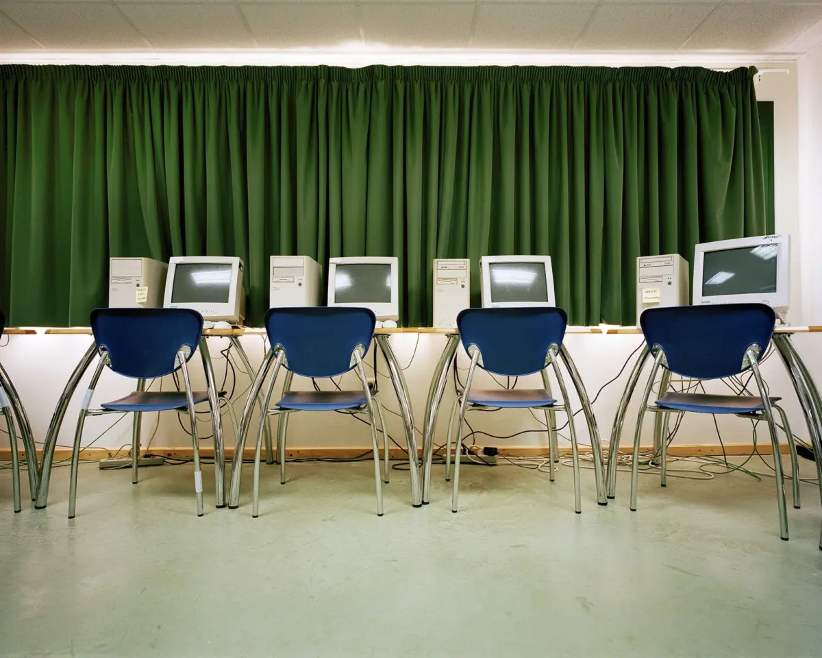 Row of beige vintage CRT computer monitors on desks in a computer lab, with green curtain backdrop and blue chairs in front.