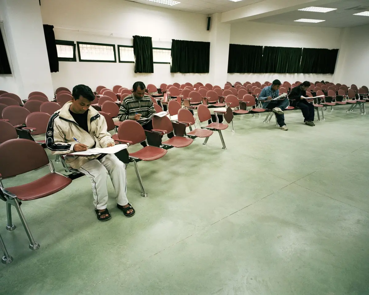 Students in a largely empty lecture hall, writing on papers; rows of pink chairs, windows with black curtains along the back.