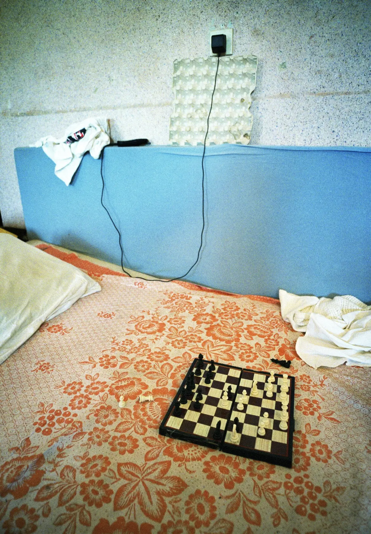 Chessboard with pieces mid-game on a floral bedspread in a cluttered bedroom.