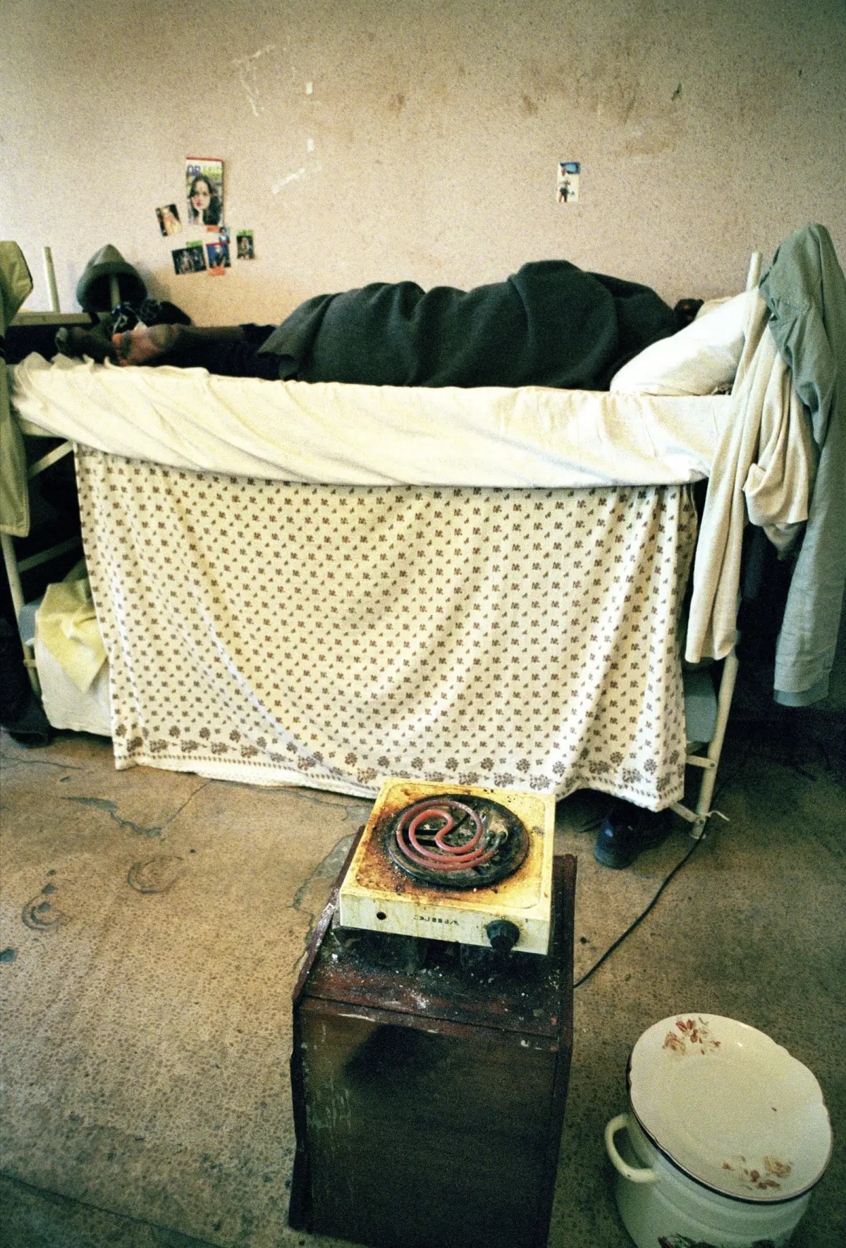 Person sleeping on a loft bed covered with dark blanket in a cluttered dorm room with posters on the wall