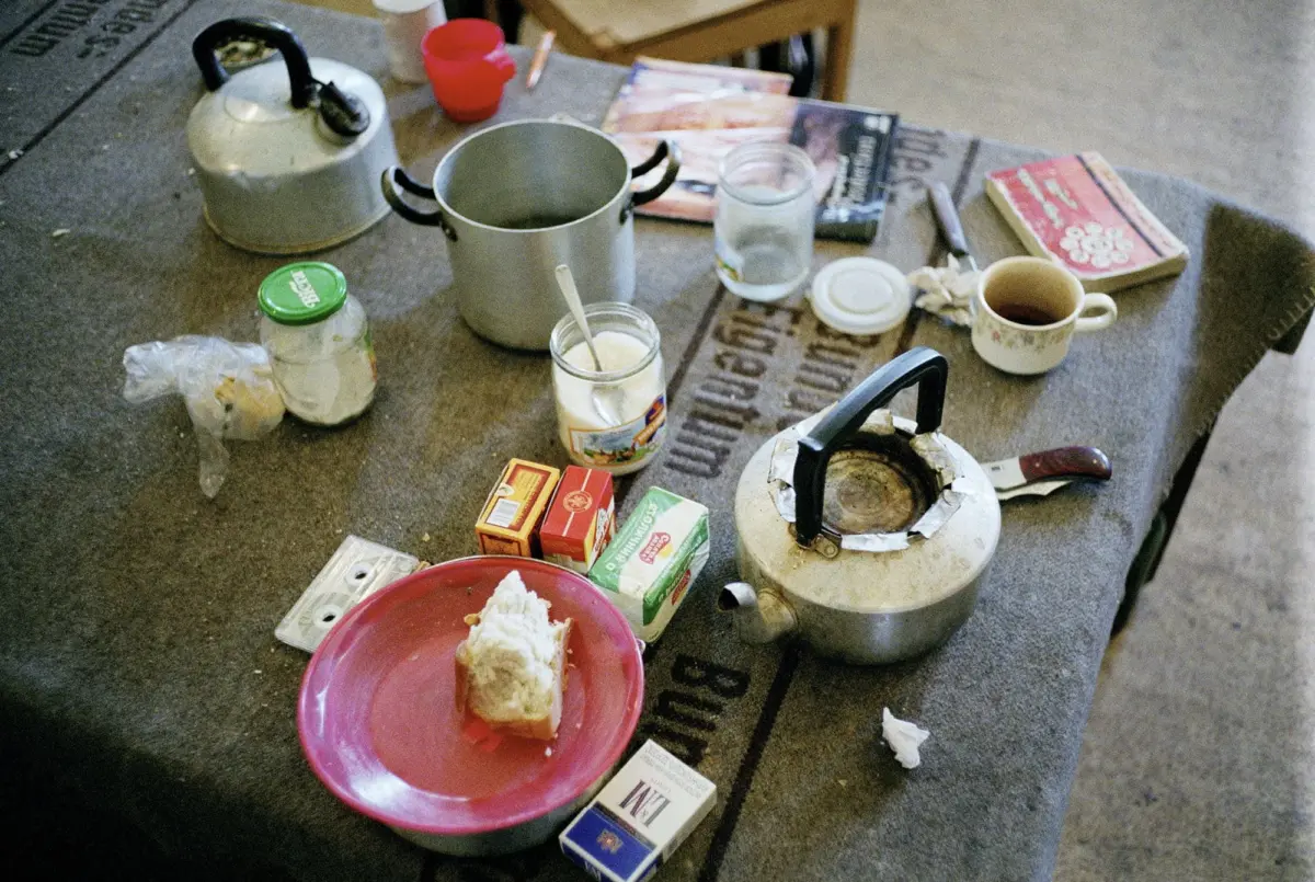 Outdoor table cluttered with kettle, metal pot, mug, glass jar, red plate with bread, butter boxes, and magazines.