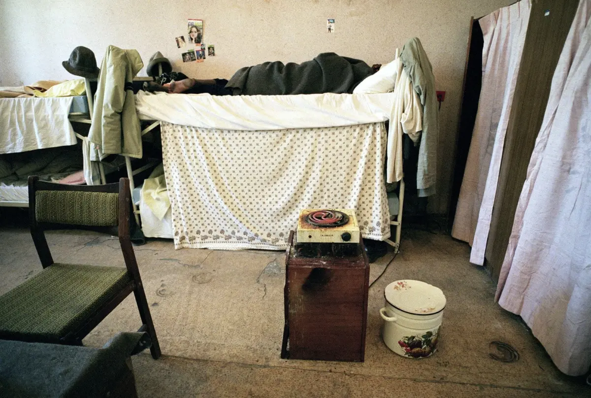 Dorm room with a top bunk and blanket-covered sleeping person, a small stovetop on a wooden stand, and a floral bucket on the floor.