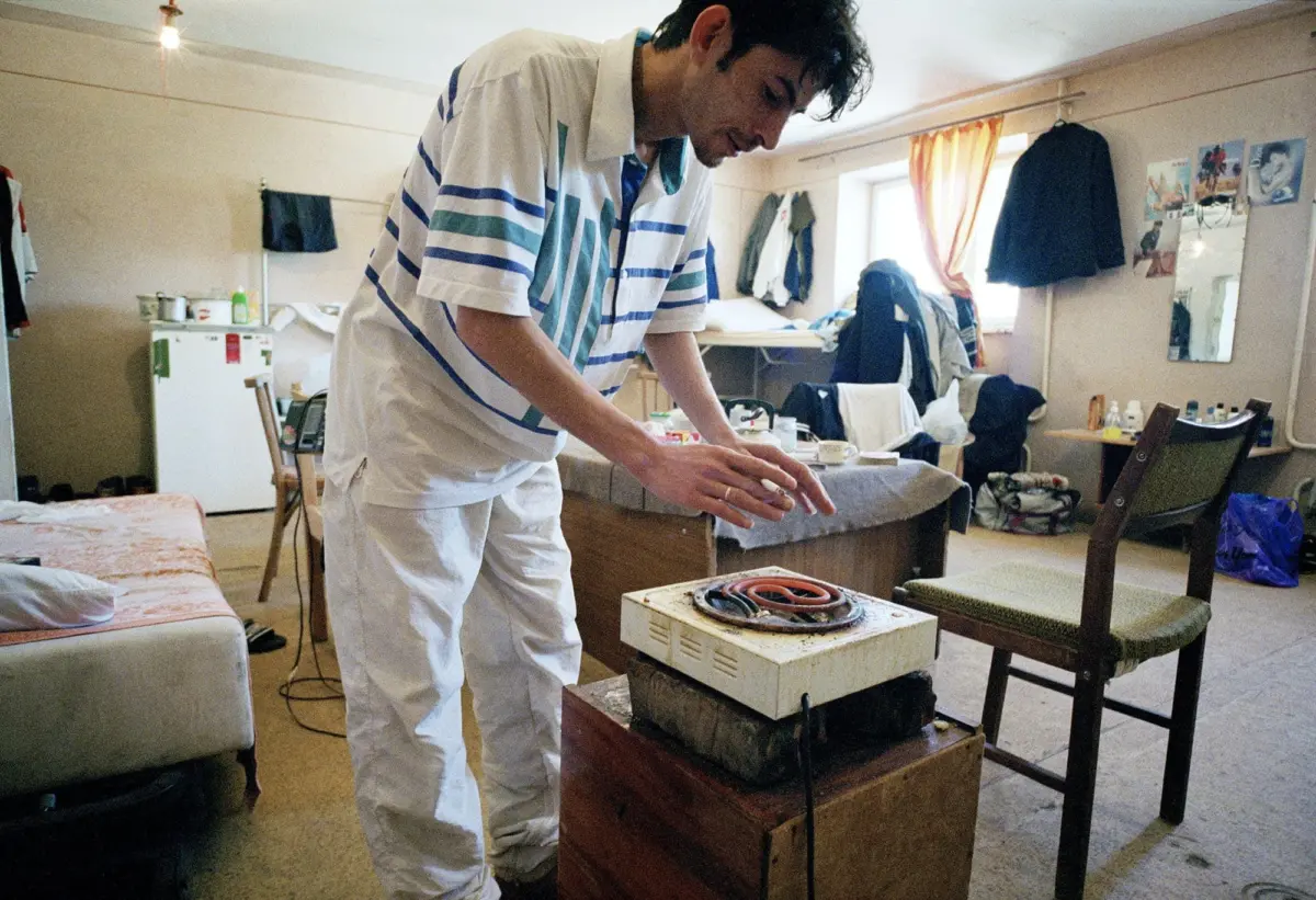 Person bends over an electric hot plate on a wooden box in a cluttered bedroom with clothes, coats, and posters.