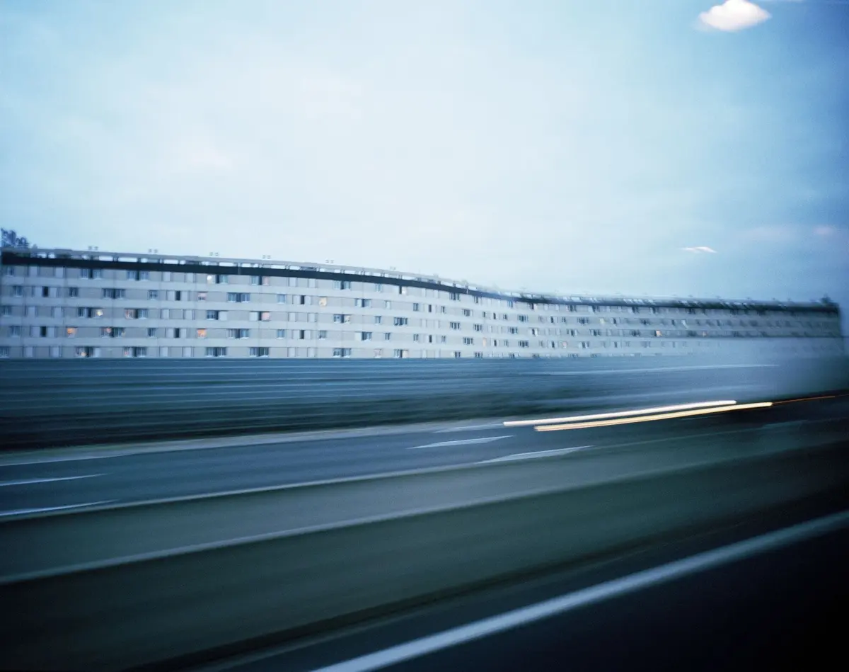Motion-blurred highway with guardrail; long row of multi-story apartment buildings along the road at dusk.