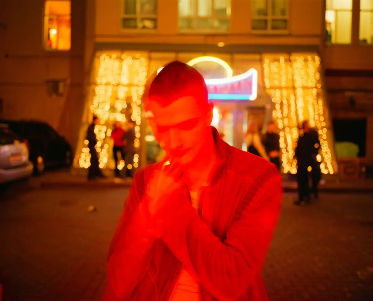 Person wearing a red jacket seen from behind on a nighttime city street, with warm storefront lights and string lights in the background.