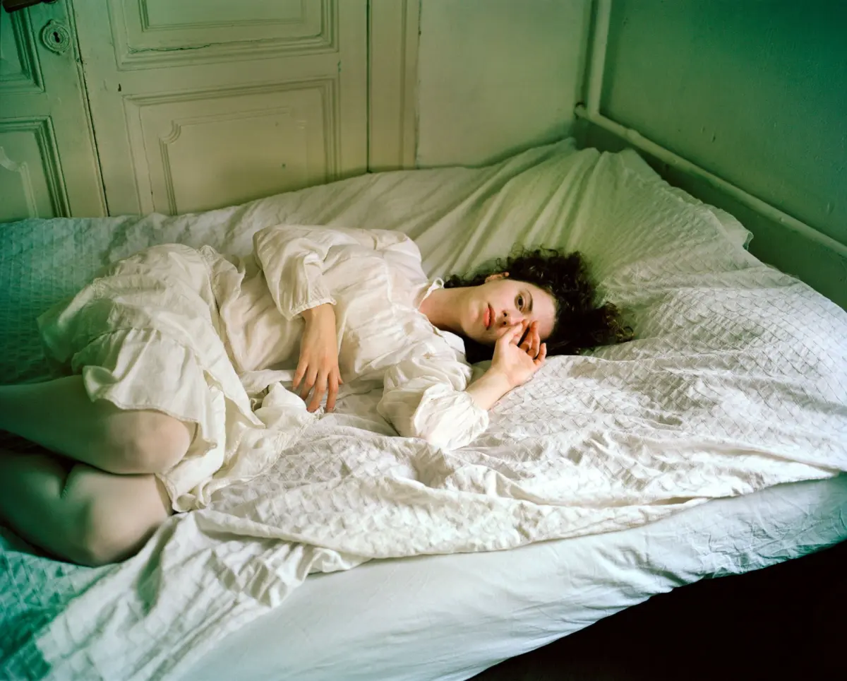 Person with curly hair lying on white bedding, wearing a white shirt, in a green-painted room with a carved door.