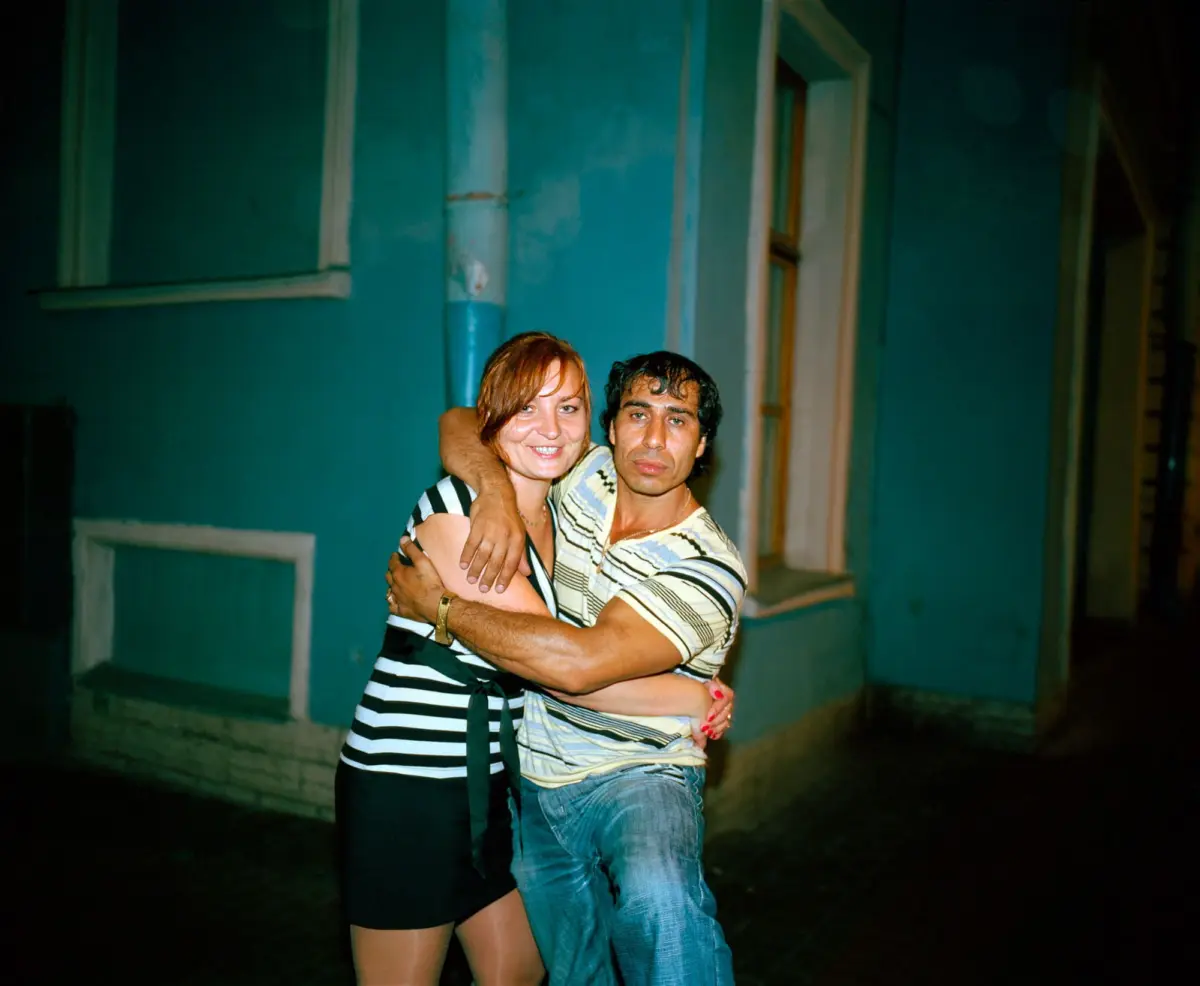 Two adults embrace in front of a teal building at night; woman in black-and-white striped dress hugs man in striped t-shirt and jeans.