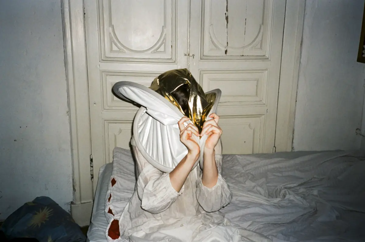 Person in white robe on a bed, lifting a gold foil hood to their face in front of ornate white doors.