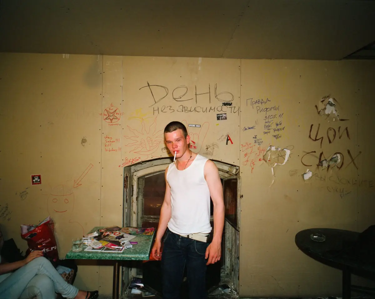 Adult person in a white tank top standing in graffiti-covered room, smoking, beside a cluttered table with magazines.