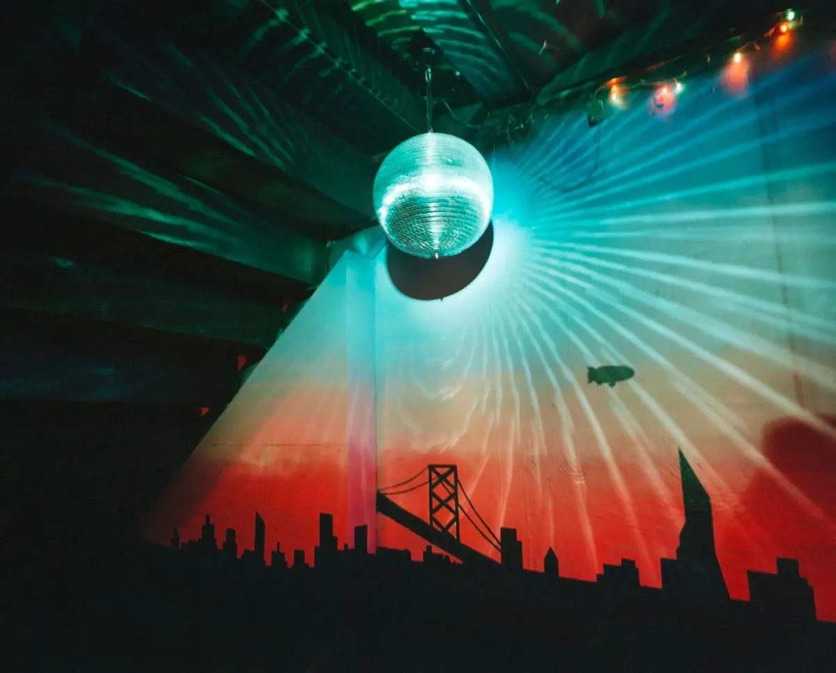 Disco ball suspended from ceiling, casting teal light beams across a wall with a city skyline mural.