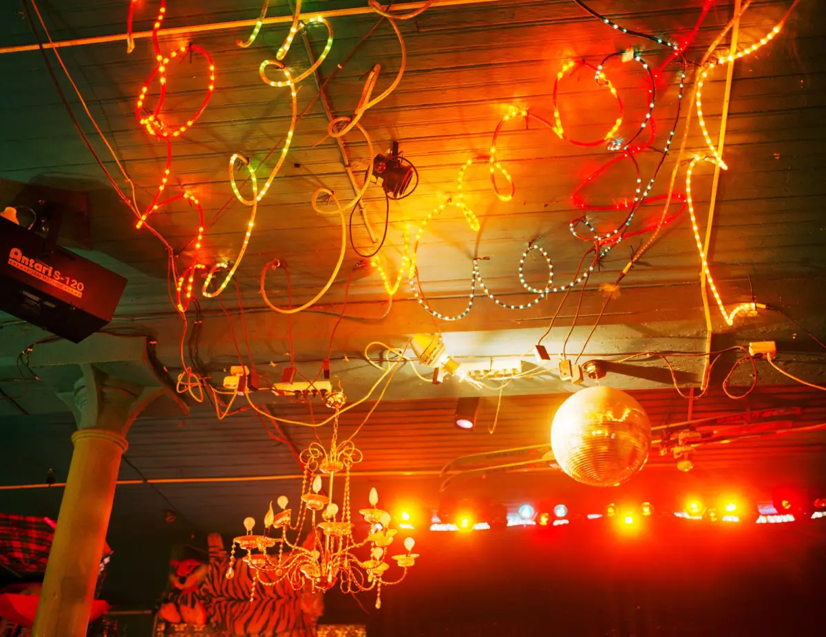 Colorful LED strands and spiraled wires hang from the ceiling over a gold chandelier and disco ball, with warm stage lights in a festive nightclub.