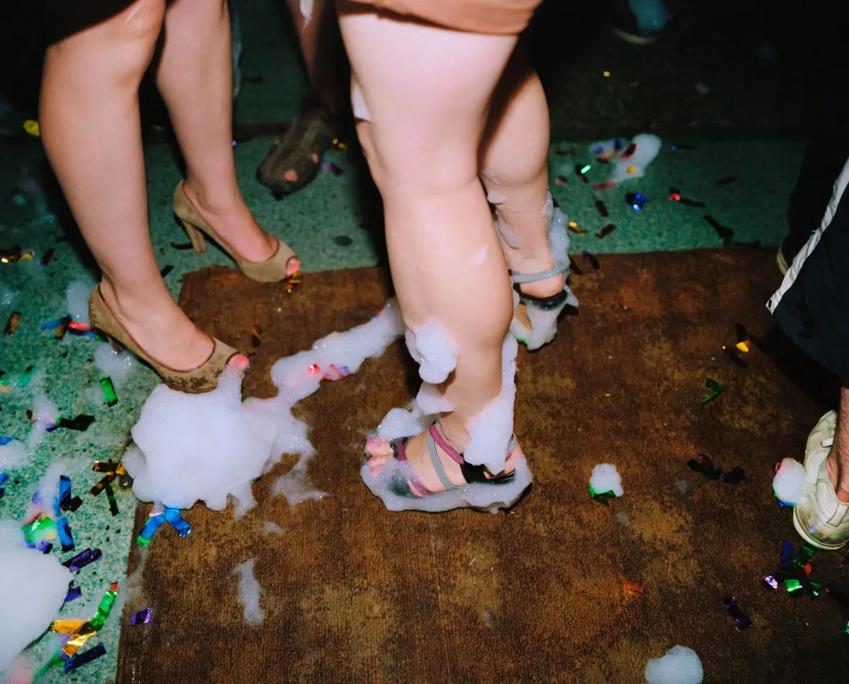 Legs of partygoers standing in foam on a carpeted floor amid confetti.
