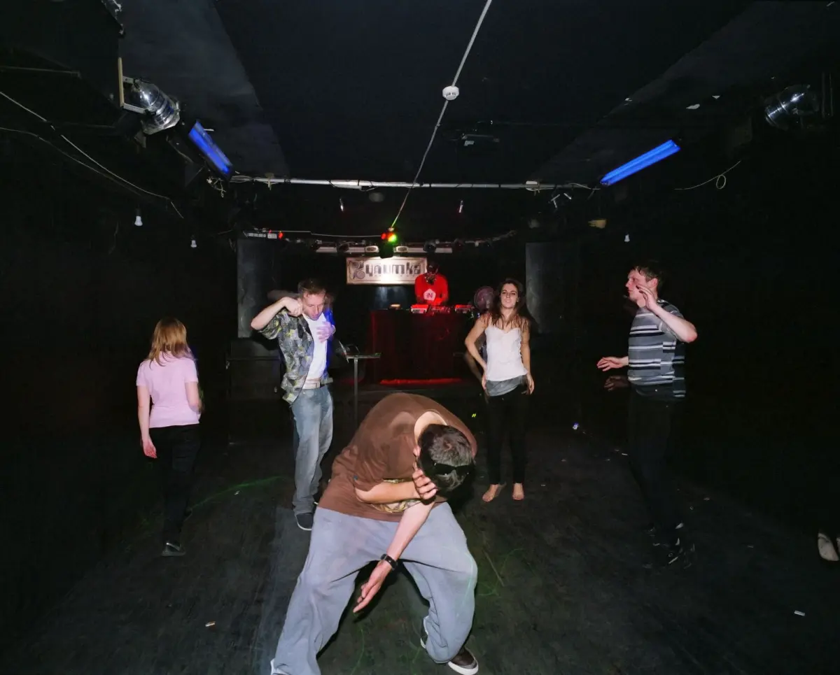 Diverse group dancing in a dim nightclub; blue tube lights and a DJ booth visible at the back.